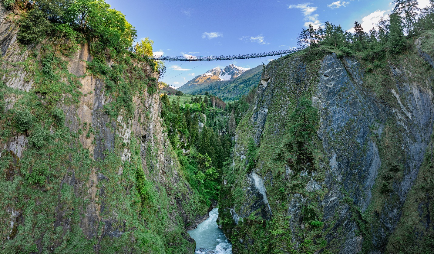 Hängebrücke über eine grüne Schlucht, dahinter ein schneebedeckter Gipfel